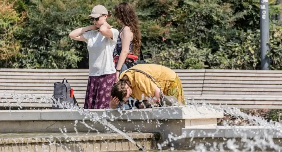 Three people stand by a splash fountain in a park; one person in a yellow shirt leans forward, head in hands, while two others watch nearby.