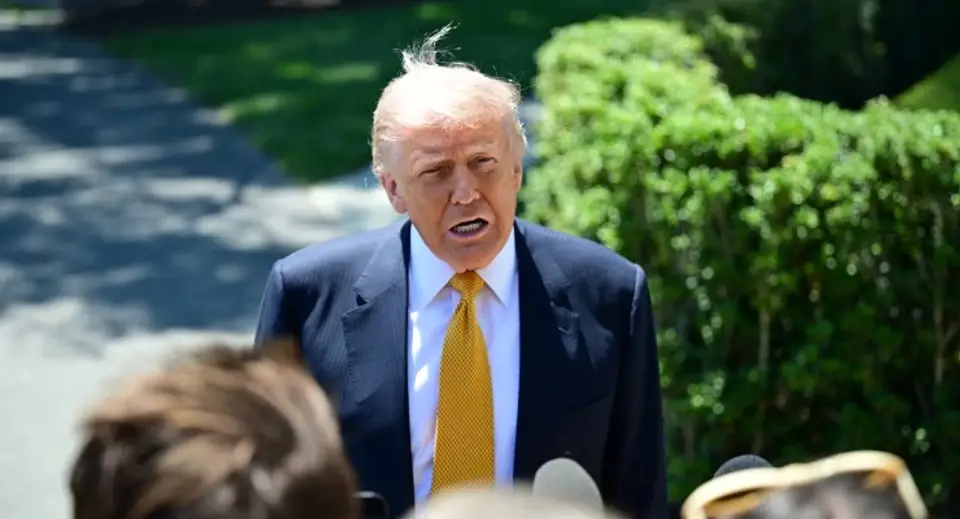 Man in a navy suit with a yellow tie speaks to reporters outdoors, microphones in front.