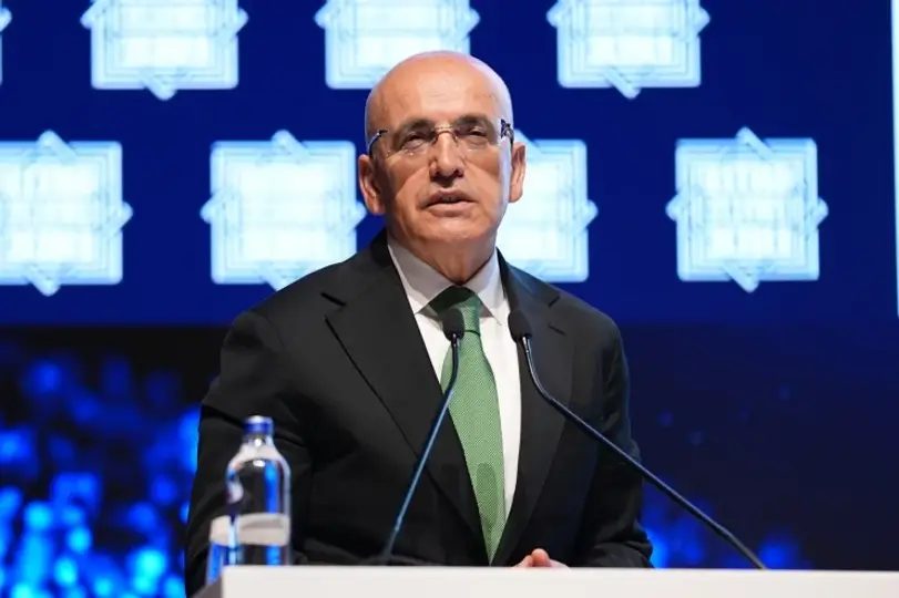 Man in a dark suit and green tie delivers a speech at a podium, water bottle nearby, blue patterned backdrop.