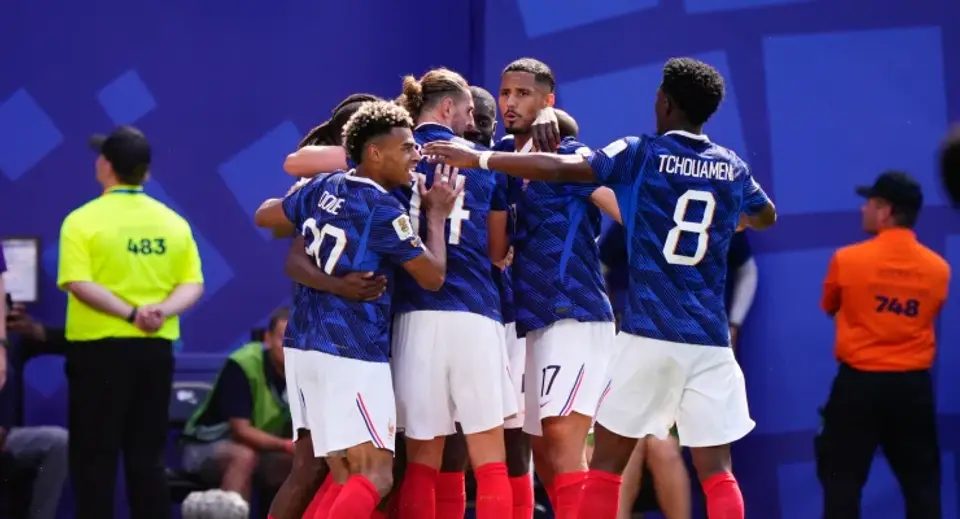 Soccer players in blue jerseys embrace in a group hug on the field, red socks visible, with team members