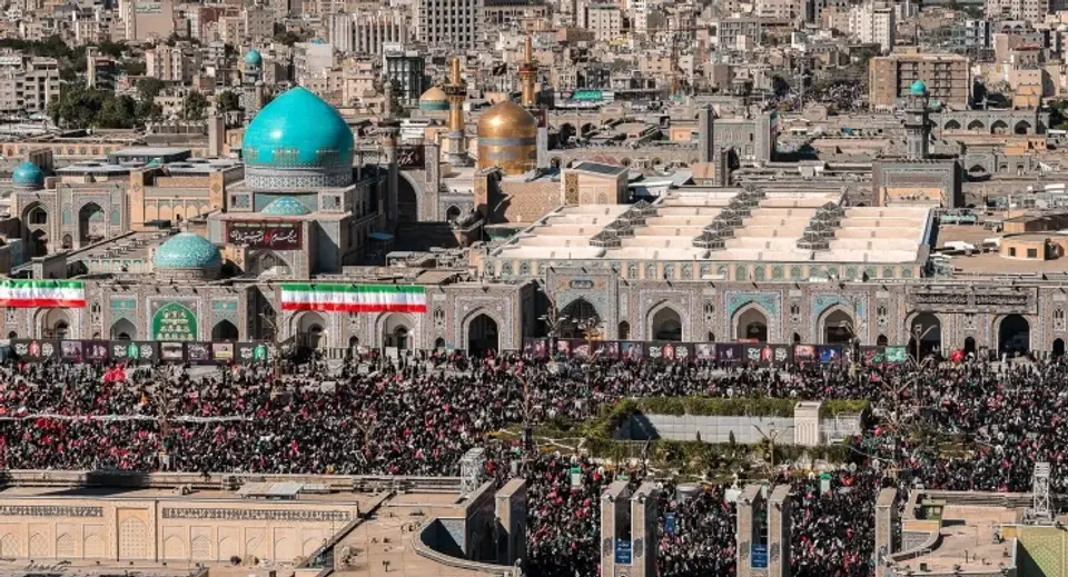 Crowded courtyard of a Persian shrine with turquoise and gold domes, expansive prayer halls, and worshippers gathered outside.