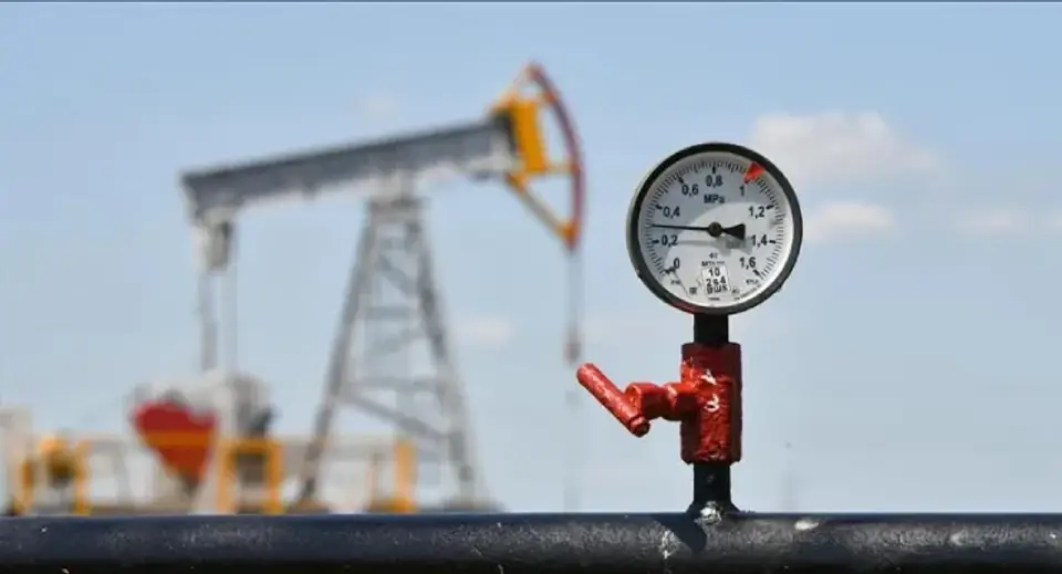 Close-up of a red valve and pressure gauge on an oil pipeline, with a pumpjack in the blurred background behind it.