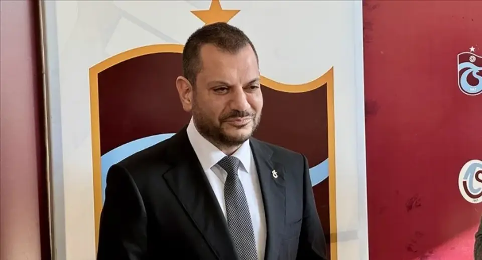 Man in a black suit and gray tie stands indoors at a formal event, with a shield crest in the background.