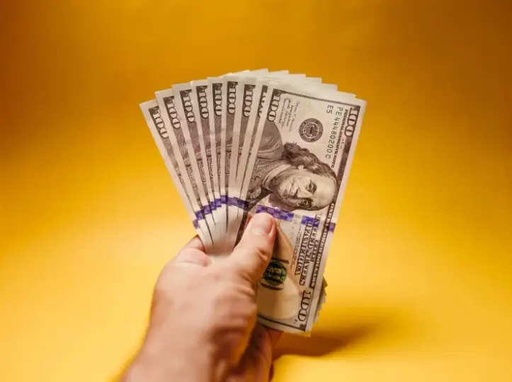 Close-up of a hand fanning multiple $100 bills against a warm yellow background, symbolizing cash.
