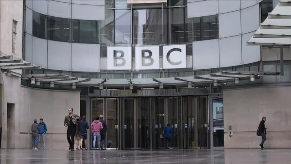 BBC headquarters entrance with large sign above glass doors and people entering the building outside the modern facade.